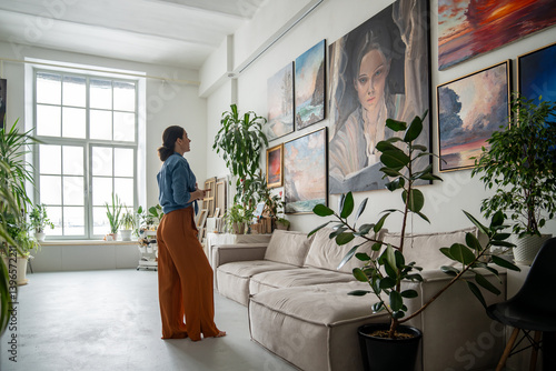 Fototapeta Naklejka Na Ścianę i Meble -  Female artist deeply reflects on portrait amidst other artworks on wall in studio. Creative workshop space with diverse collection of paintings, woman painter inspiration and new ideas for work