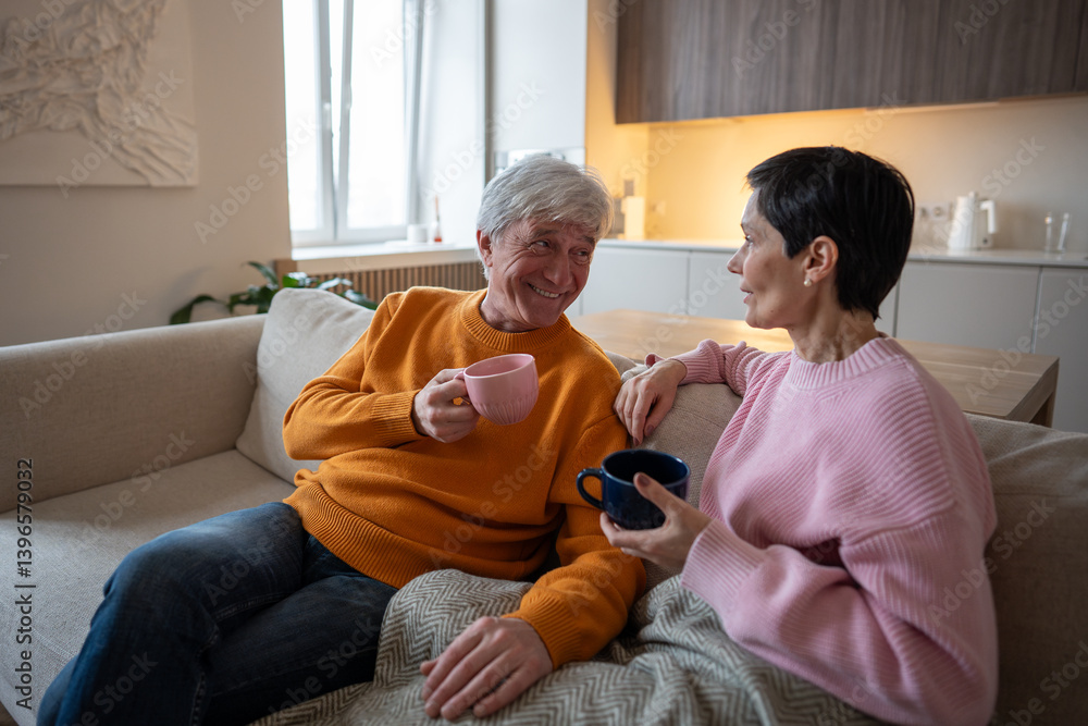 Obraz premium Joyful senior couple of husband and wife share laughter and stories, relaxing on comfortable couch with hot tea. Warm companionship, romantic hygge rest, glad smiling spouses during tea time at home