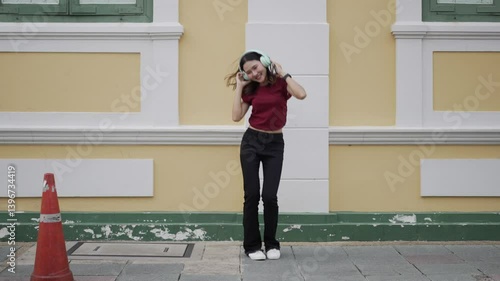 Young Asian woman wearing wireless headphones smiling while walking and listening to music on sidewalk, relaxed, stylish, and natural moment in urban area