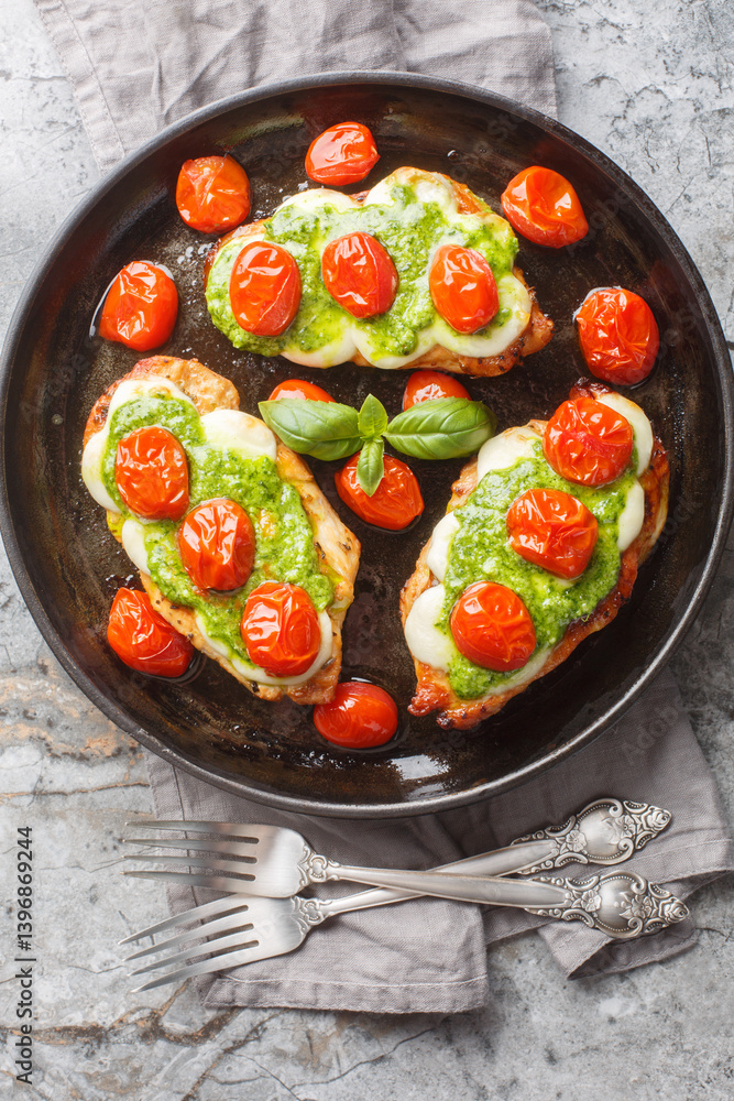 Margherita chicken that features pan-fried chicken breasts topped with melted mozzarella, pesto and tomatoes close-up on pan on table. Vertical top view from above