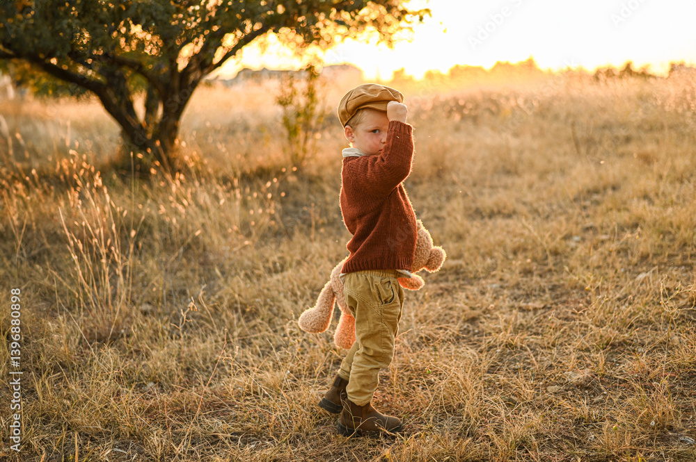 Little boy with a teddy bear walking through an olive grove at sunset. Warm rural scene with yellow grass, soft light, and peaceful childhood atmosphere. Little boy with a teddy bear walking through an olive grove at sunset. Warm rural scene with yellow grass, soft light, and peaceful childhood atmosphere.