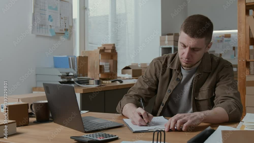 Caucasian young male storage room employee working on laptop and writing data down on piece of paper at wooden table during workday Caucasian young male storage room employee working on laptop and writing data down on piece of paper at wooden table during workday