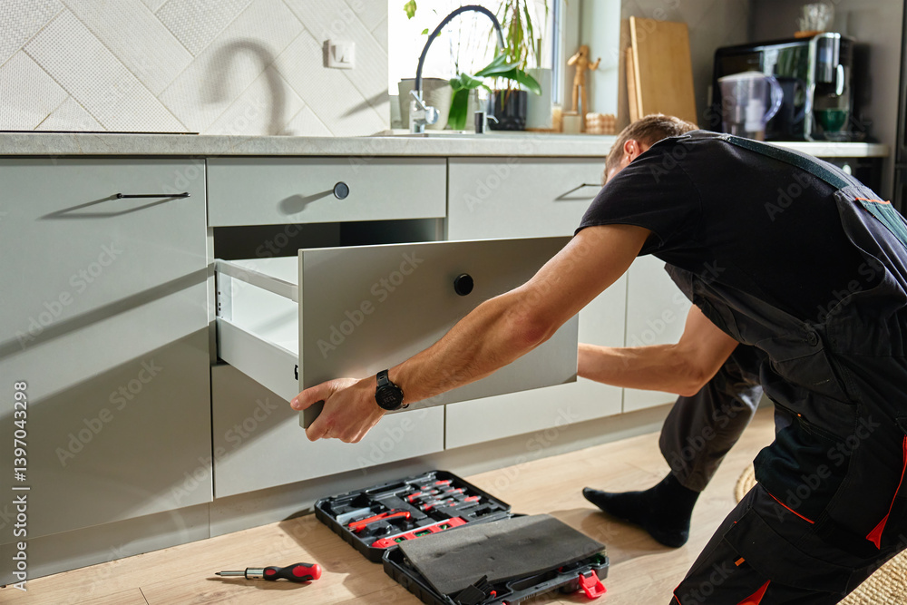 Man assembling kitchen drawer in modern cabinet using screwdriver. Handyman installing and adjusting drawer slide mechanism in contemporary kitchen. Furniture installation