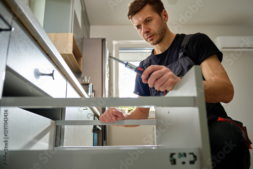 Obraz na płótnie Man assembling kitchen drawer in modern cabinet using screwdriver. Handyman installing and adjusting drawer slide mechanism in contemporary kitchen. Furniture installation w salonie