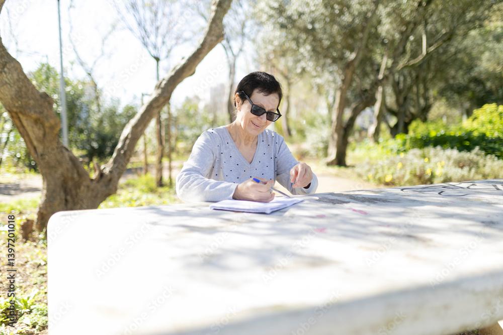 Fototapeta premium Senior woman writing in notebook at park table on sunny day