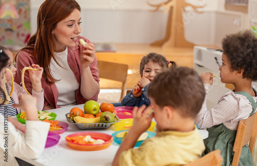A diverse group of preschool children sits around a table eating healthy snacks while a teacher engages with them, promoting nutrition, inclusion, and social interaction in early education.