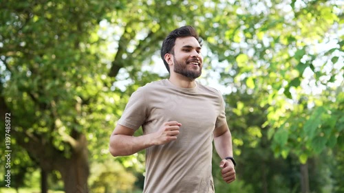 Handsome male runner jogging in an urban city park and looking smart watch. Young athlete using fitness tracker. Sportsman checking result. Adult fit man in T-shirt enjoys running outdoors in nature