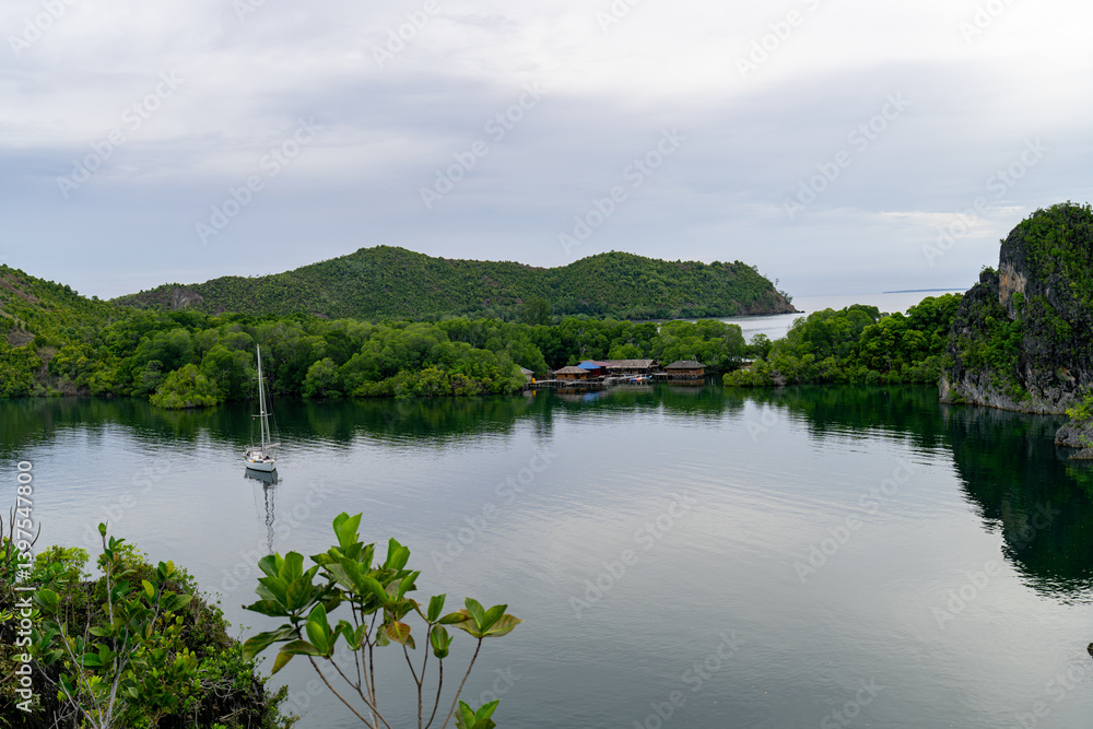 Fototapeta premium Calm bay with sailboat anchored near a village surrounded by forested hills