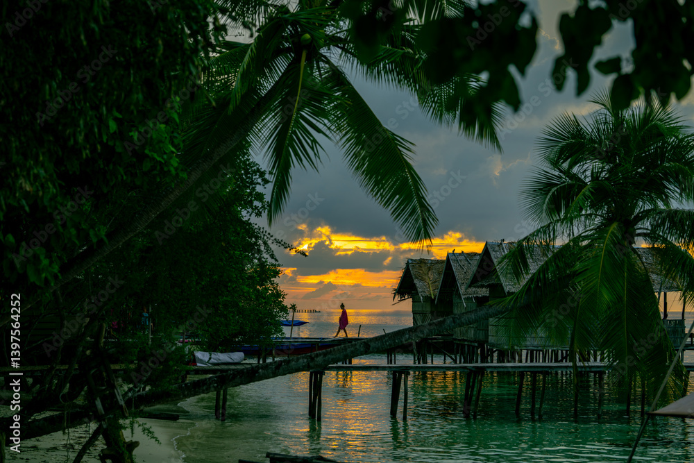 Fototapeta premium Man walking on wooden bridge above calm tropical water with glowing sunset in background