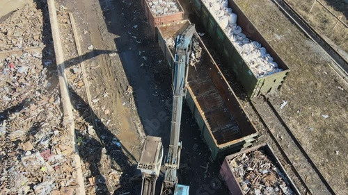 An aerial view captures a large crane lifting metal scraps in an industrial site. Surrounding debris and containers indicate the aftermath of material processing.