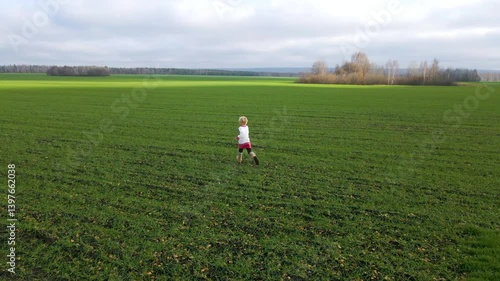 A young child joyfully explores a wide green field surrounded by distant trees. The sky is overcast, reflecting a serene autumn day. The scene captures a sense of adventure and freedom