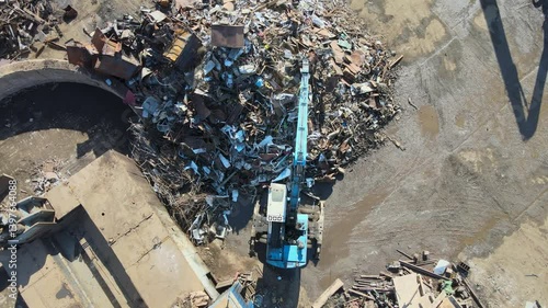 An aerial view captures a large crane lifting metal scraps in an industrial site. Surrounding debris and containers indicate the aftermath of material processing.