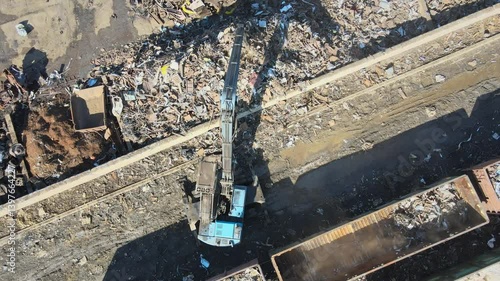 An aerial view captures a large crane lifting metal scraps in an industrial site. Surrounding debris and containers indicate the aftermath of material processing.