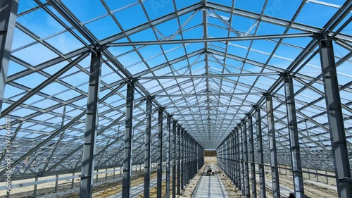Flight inside the large metal building structure is taking shape in an agricultural zone. Metal frames are installed, creating an expansive space for future cultivation growth or animal husbandry