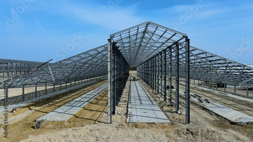 Flight past the large metal building structure is taking shape in an agricultural zone. Metal frames are installed, creating an expansive space for future cultivation growth or animal husbandry