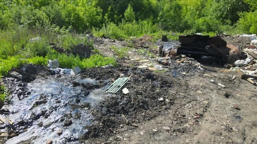 An abandoned industrial site shows waste and debris scattered amid overgrown vegetation. The polluted bitumen area highlights the impact of neglect on the environment