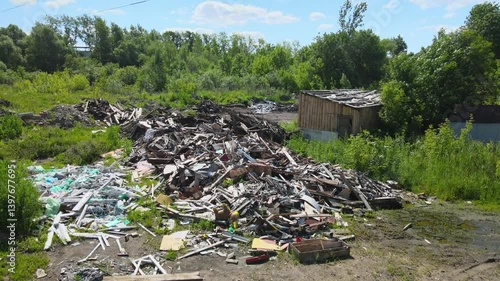 An abandoned lot features a large pile of debris, including wood and scrap materials, surrounded by lush green vegetation and trees. The setting reflects neglect and natural reclamation