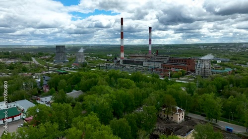 A large energy production facility stands prominently amidst lush green trees under a cloudy sky. Smoke rises from the stacks, indicating active operations in the area.
