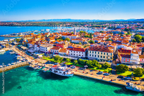 Aerial view of the vibrant waterfront promenade in Biograd na Moru, Dalmatia, Croatia, featuring a lively marina, moored boats, traditional Mediterranean buildings, and the shimmering blue waters