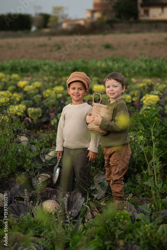 Two young boys in vintage-style clothes stand in a cabbage field with a basket and a small shovel, looking at a cabbage head, evoking rural childhood and simple farm life.