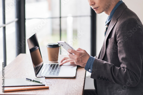 ノートパソコンを開き、スマートフォンを見ながら作業をしている日本人男性。