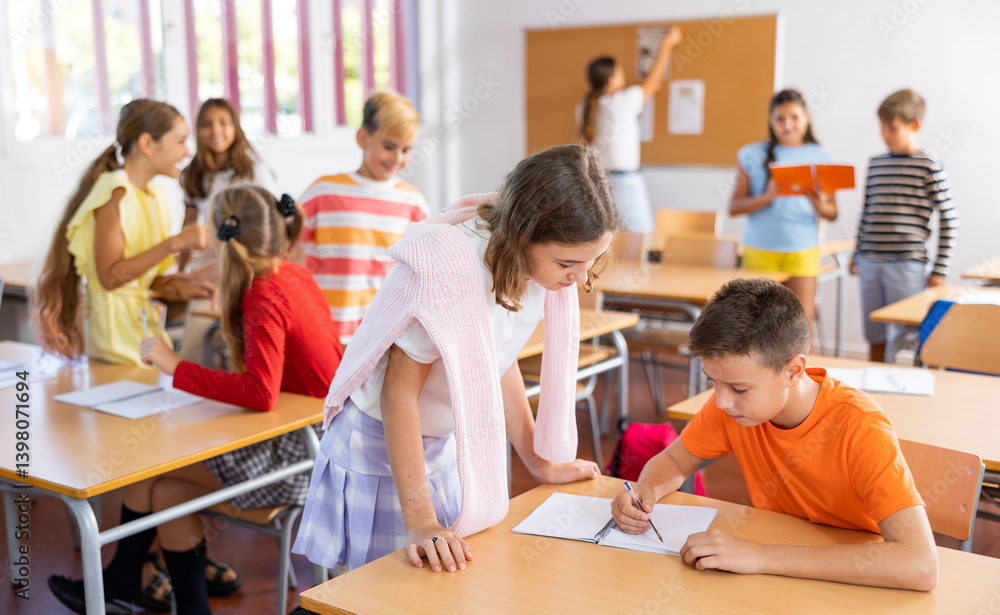 Obraz premium Positive friendly tween girl helping classmate sitting at school desk to complete written assignment in notebook during break before lesson ..