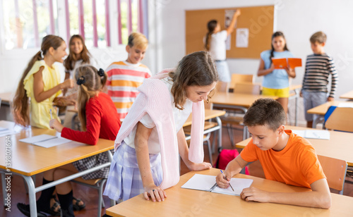 Positive friendly tween girl helping classmate sitting at school desk to complete written assignment in notebook during break before lesson ..