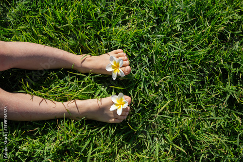 Fototapeta Naklejka Na Ścianę i Meble -  child bare feet with plumeria flowers between toes on green lawn . joy, cheerful positive atmosphere, happy childhood, relaxation. Hello summer. energy of nature. Earth day
