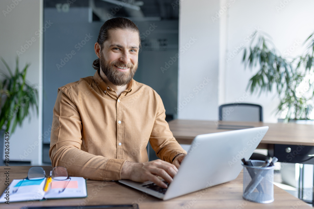 A bearded man smiles at the camera while working on a laptop. The office environment shows the focus on productivity and work.