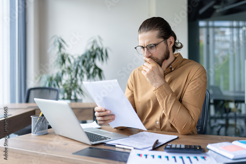 A focused professional reviews documents at his desk in an office setting, pondering the information. Laptop, papers, and calculator are visible.