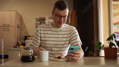 Young man enjoying morning routine, sitting at kitchen table while eating pancakes and sipping coffee, simultaneously browsing smartphone with relaxed demeanor