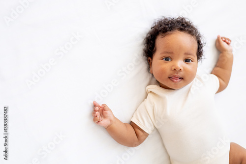 close-up portrait of a small African American baby girl in a white bodysuit on a cotton bed at home, funny six-month-old smiling joyful black newborn baby shows tongue