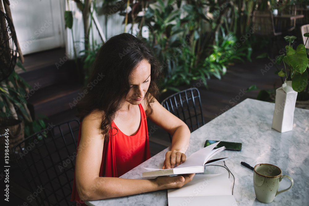 Woman in a red dress reading a book at a marble table in a lush, plant-filled environment. Coffee mug, notebook, and smartphone on table suggest a relaxed, focused atmosphere of study or reflection