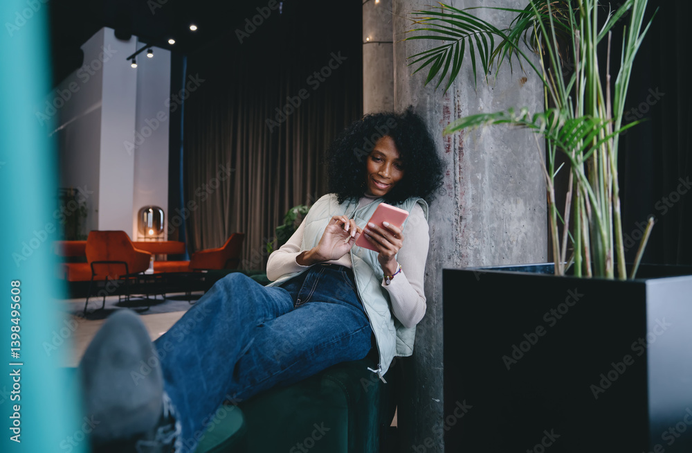 Fototapeta premium Stylish woman scrolling on her cellphone while booking hotel or flight. Dark skinned female enjoys convenience of modern technology, planning her next trip with few taps on screen of mobile phone