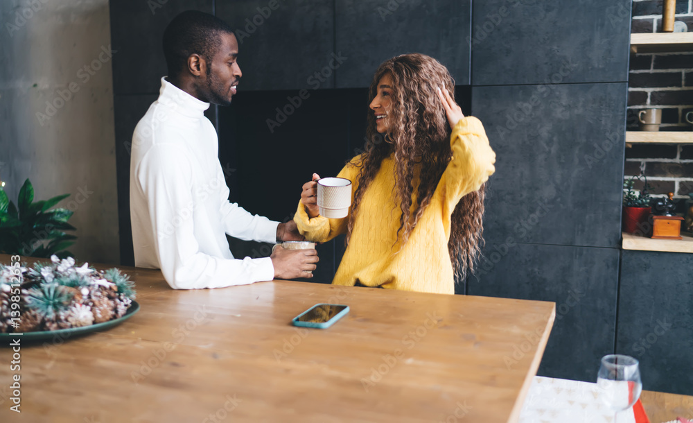 Young Black woman in a yellow sweater and Black man in a white turtleneck sharing a joyful moment with coffee cups in a modern kitchen setting, conveying warmth and happiness