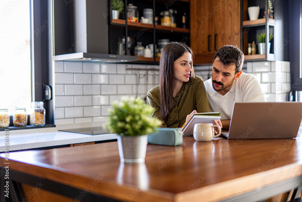 Young couple calculating domestic finances using laptop and notebook, managing their budget and expenses in their modern kitchen Young couple calculating domestic finances using laptop and notebook, managing their budget and expenses in their modern kitchen