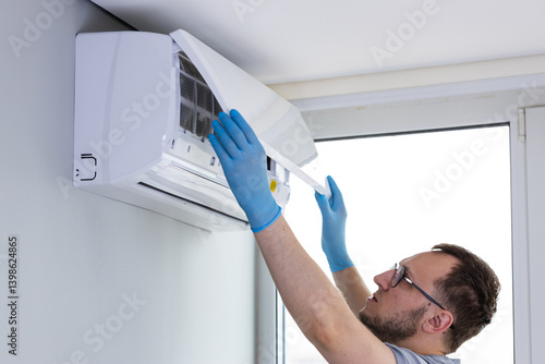 A Technician Performing Maintenance on a Wall-Mounted Air Conditioner Unit in a Bright Room with Large Windows, Wearing Blue Gloves and Focusing on Cleaning the Filter