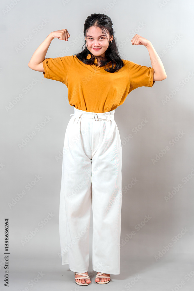 Smiling young Asian woman proudly flexing both arms in a strong pose, exuding confidence, charm, and determination—ideal for themes of empowerment, positivity, and motivation. Smiling young Asian woman proudly flexing both arms in a strong pose, exuding confidence, charm, and determination—ideal for themes of empowerment, positivity, and motivation.