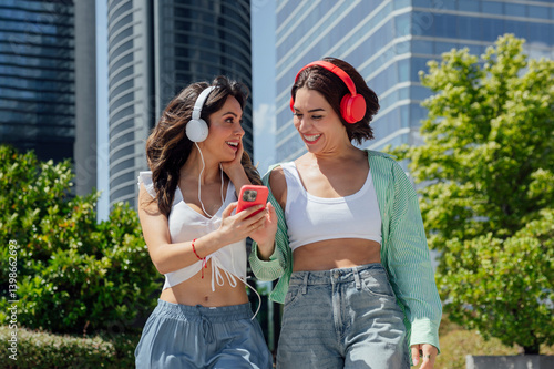 Two young women sharing earphones listening to music on a smartphone in the city