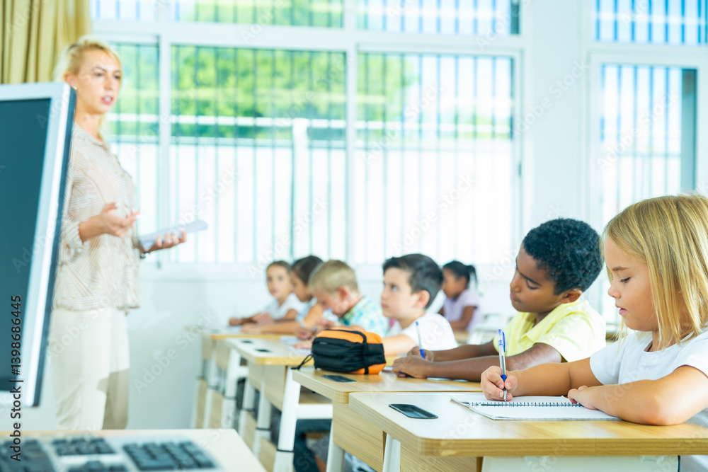 Focused preteen pupils studying in classroom with female teacher, writing exercises in workbooks Focused preteen pupils studying in classroom with female teacher, writing exercises in workbooks