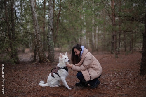 A woman in a beige coat gently pets her white dog in a harness while crouching in a forest. The scene captures a peaceful moment with pine trees in the background, showcasing a warm, loving bond.