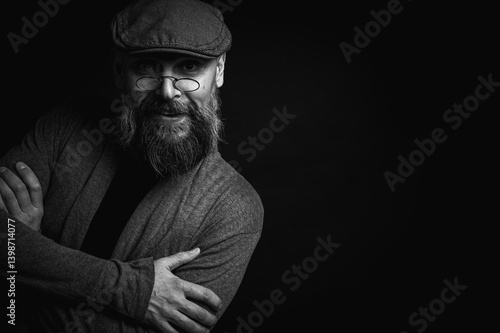 Obraz na płótnie Black and white, portrait of an emotional bearded man in a gray hat, gray sweater and glasses looking at the camera on a dark background. Dramatic portrait in dim lighting, expressive face, emotional w salonie
