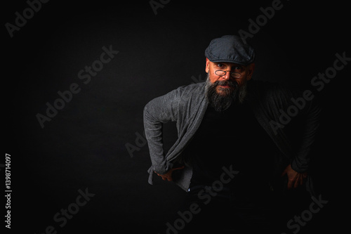 Obraz na płótnie Portrait of an emotional bearded man in a gray hat, gray sweater and glasses looking at the camera on a dark background. Dramatic portrait in dim lighting, expressive face, emotional w salonie