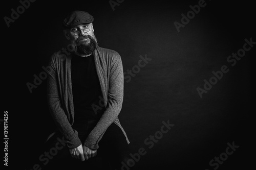 Obraz na płótnie Black and white, portrait of an emotional bearded man in a gray hat, gray sweater and glasses looking at the camera on a dark background. Dramatic portrait in dim lighting, expressive face, emotional w salonie