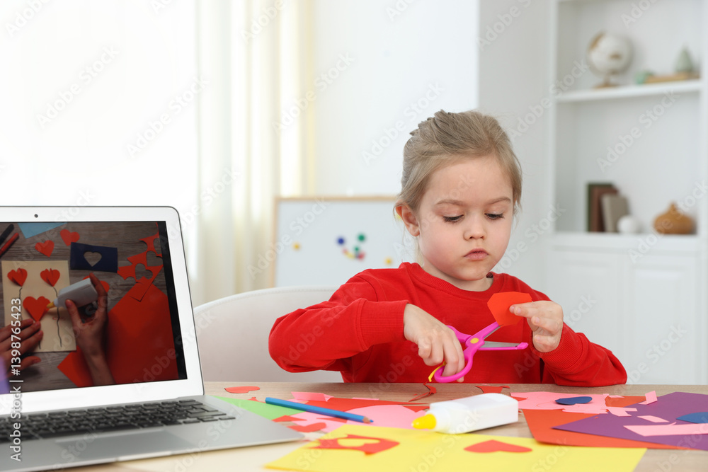 Little girl making handmade project with color paper and scissors by video tutorial at table indoors Little girl making handmade project with color paper and scissors by video tutorial at table indoors
