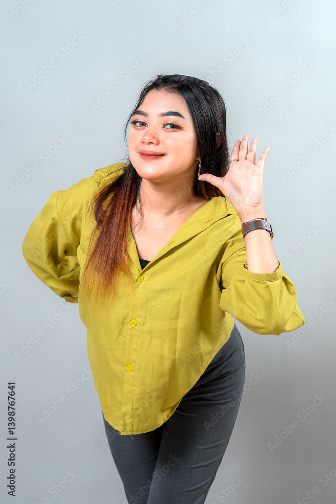 Fototapeta premium Young woman with a curious facial expression, smiling while cupping her ear to listen closely. A playful and engaging gesture perfect for themes of attention, hearing, or communication.