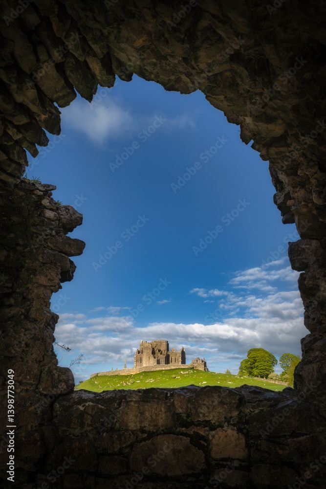 Picturesque view of ancient historic ruins of Rock of Cashel, framed beautifully by stone arch of Hore Abbey, with striking clear blue skies and rolling green hills in the distance, Tipperary, Ireland