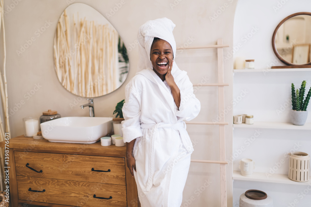 Beautiful positive moment of pure happiness during morning routine. Portrait of cheerful dark skinned woman in white bathrobe and towel expressing genuine joy and expression in bathroom