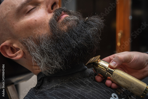 Obraz na płótnie Close-up of a barber trimming a beard with electric clippers in a modern barbershop. Professional men's grooming and beard care service, selective focus w salonie