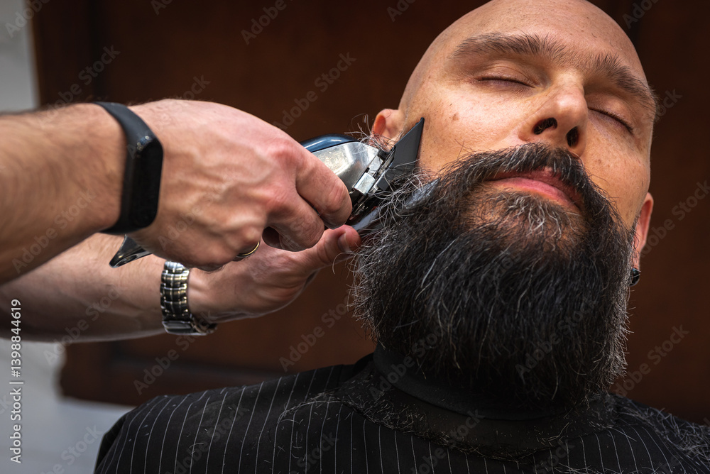 Close-up of a barber trimming a beard with electric clippers in a modern barbershop. Professional men's grooming and beard care service, selective focus
